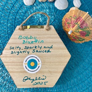 May include: Wooden, hexagonal sign with the text "Bobby Bluefin, salty, sparkly and slightly sauced." The sign has a jute rope handle and a small sticker that says "Sunshine Utopia." A decorative cocktail umbrella and seashells are in the background.