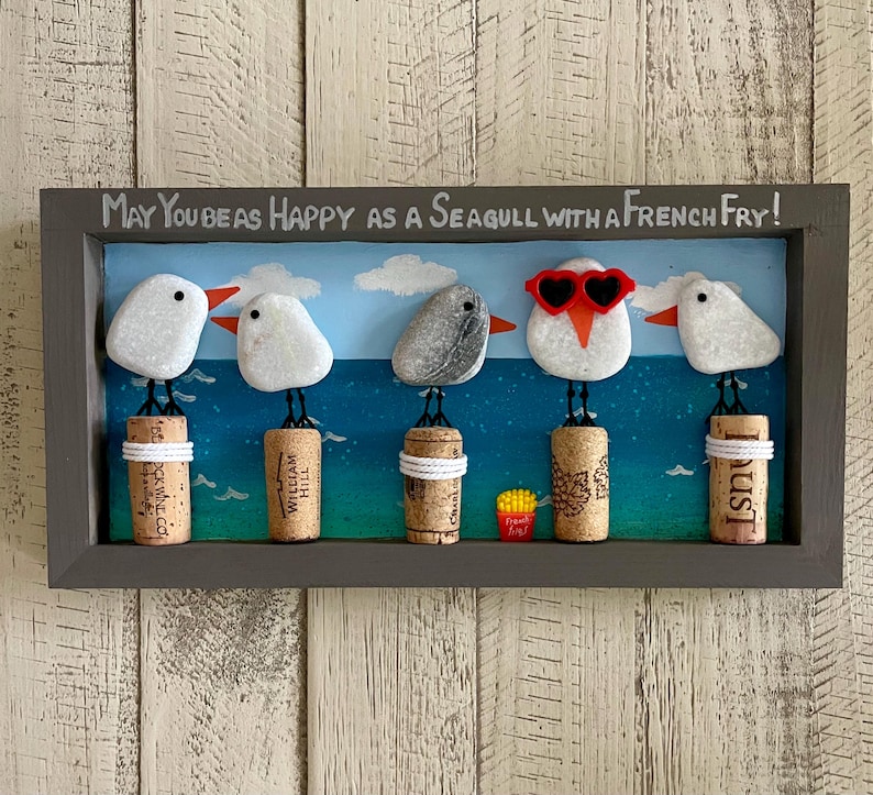 May include: A rectangular wall art piece in a dark gray frame featuring five pebble seagulls perched on wine corks against a painted blue ocean background. The text reads, "May you be as happy as a seagull with a french fry!" One seagull wears red heart-shaped sunglasses.