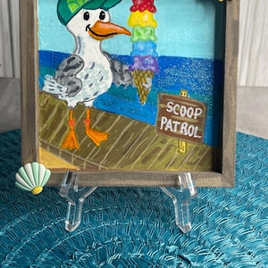 May include: A framed painting depicts a seagull wearing a green cap, holding a colorful ice cream cone. The artwork features a blue ocean backdrop and a sign that reads "SCOOP PATROL". A yellow sun charm is attached to the frame.