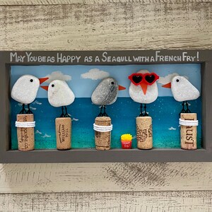 May include: A rectangular wall art piece featuring five pebble seagulls perched on wine corks against a painted blue ocean background. The text reads, May You Be As Happy As A Seagull With A French Fry! One seagull wears red heart-shaped sunglasses.
