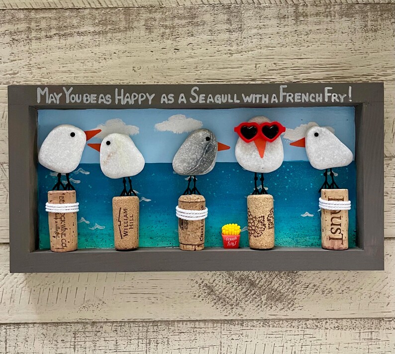 May include: A rectangular wall art piece featuring five pebble seagulls perched on wine corks against a painted blue ocean background. The text reads, May You Be As Happy As A Seagull With A French Fry! One seagull wears red heart-shaped sunglasses.