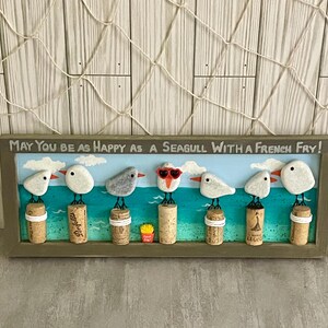 May include: A rectangular framed wall art piece featuring seven pebble seagulls perched on wine corks against a painted blue ocean background. The text reads, May You Be As Happy As A Seagull With A French Fry! One seagull wears red heart-shaped glasses.
