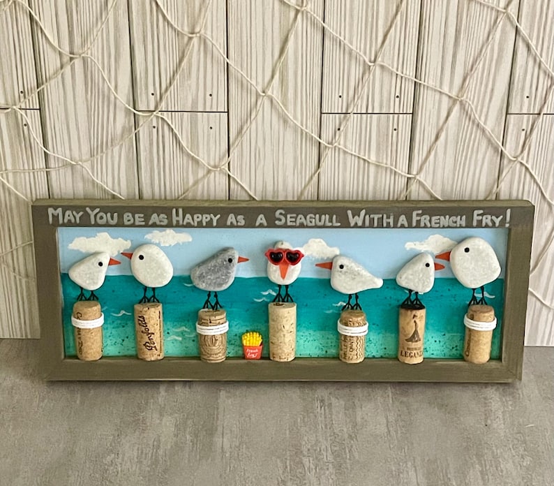 May include: A rectangular framed wall art piece featuring seven pebble seagulls perched on wine corks against a painted blue ocean background. The text reads, May You Be As Happy As A Seagull With A French Fry! One seagull wears red heart-shaped glasses.