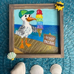 May include: A framed painting of a seagull wearing a green cap, holding a rainbow ice cream cone. The sign reads "SCOOP PATROL". The painting is surrounded by seashells and a yellow sun-shaped embellishment.
