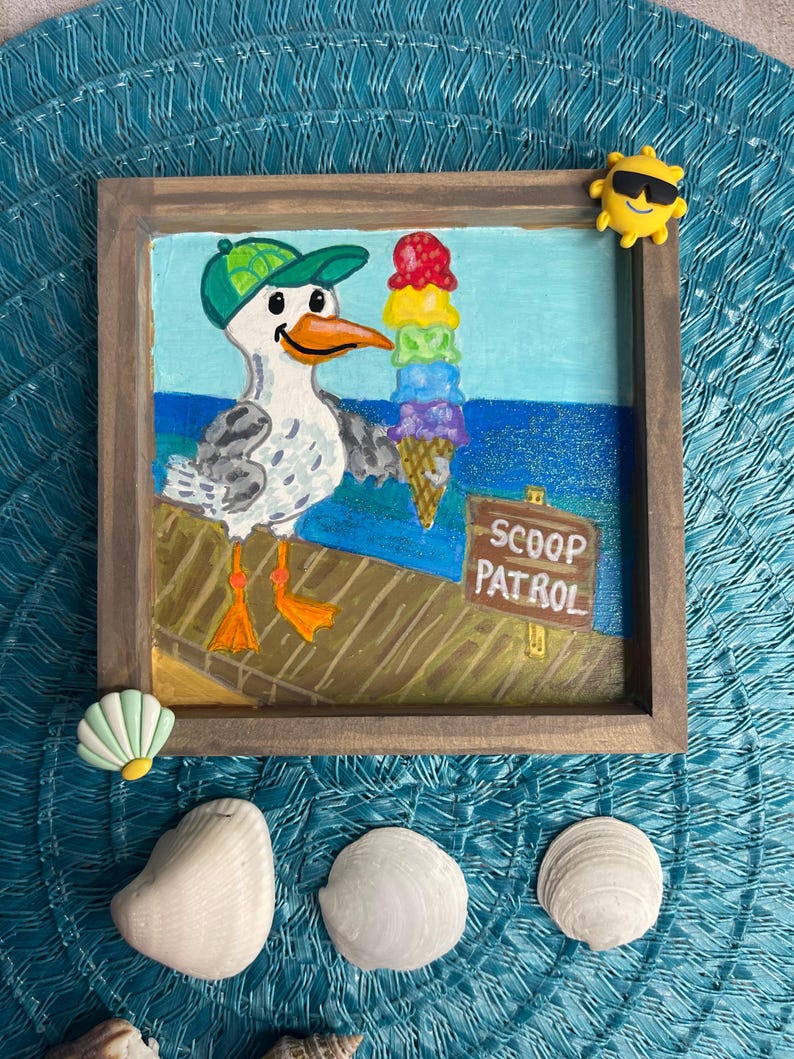 May include: A framed painting of a seagull wearing a green cap, holding a rainbow ice cream cone. The sign reads "SCOOP PATROL". The painting is surrounded by seashells and a yellow sun-shaped embellishment.