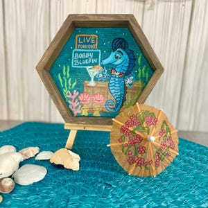 May include: A hexagonal wooden framed artwork featuring a blue seahorse in a tuxedo holding a cocktail. The artwork has the text "LIVE TONIGHT" and "BOBBY BLUEFIN". A small cocktail umbrella with pink flowers sits in front of the artwork.