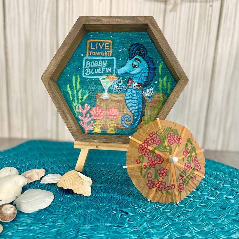 May include: A hexagonal wooden framed artwork featuring a blue seahorse in a tuxedo holding a cocktail. The artwork has the text "LIVE TONIGHT" and "BOBBY BLUEFIN". A small cocktail umbrella with pink flowers sits in front of the artwork.