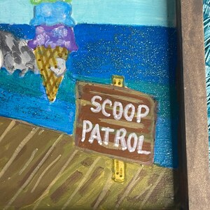 May include: A framed painting featuring a colorful ice cream cone with four scoops and a sign that reads "SCOOP PATROL." The background depicts a blue ocean and a wooden dock. The artwork is framed in a brown wooden frame.