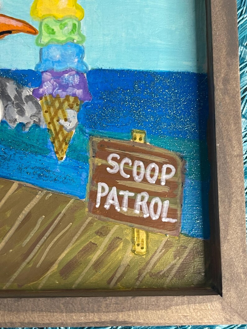 May include: A framed painting featuring a colorful ice cream cone with four scoops and a sign that reads "SCOOP PATROL." The background depicts a blue ocean and a wooden dock. The artwork is framed in a brown wooden frame.