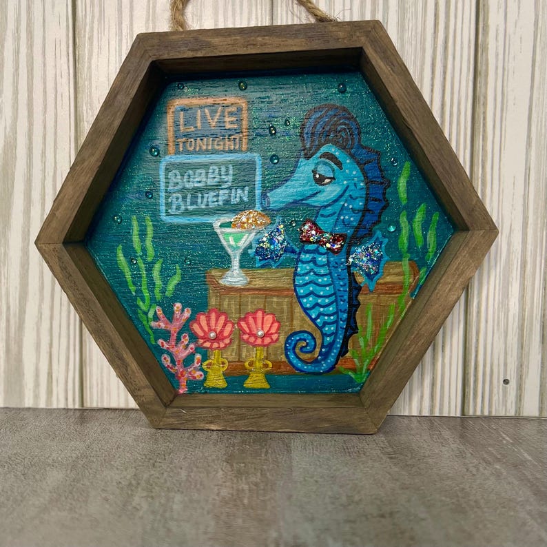 May include: A hexagonal wooden wall art piece featuring a blue seahorse holding a martini. The seahorse is in front of a bar with the text "LIVE TONIGHT BOBBY BLUEFIN". The artwork has a dark teal background with coral and seaweed.