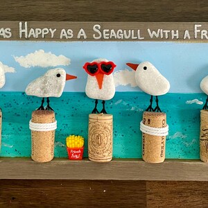 May include: A rectangular wooden framed wall art piece featuring seven white pebble seagulls perched on wine corks against a painted blue sea and sky background. The text reads, May You be as Happy as a Seagull with a French Fry! One seagull wears red heart-shaped glasses.