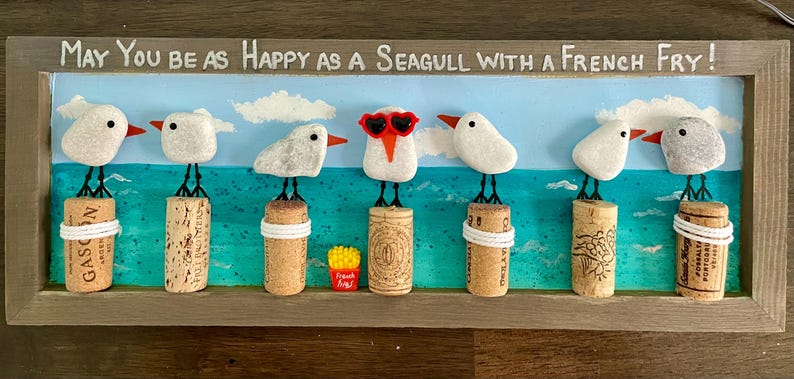 May include: A rectangular wooden framed wall art piece featuring seven white pebble seagulls perched on wine corks against a painted blue sea and sky background. The text reads, May You be as Happy as a Seagull with a French Fry! One seagull wears red heart-shaped glasses.