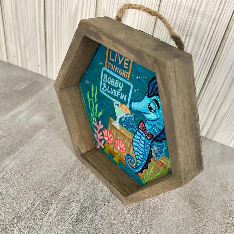 May include: A wooden, hexagonal-shaped wall hanging featuring a cartoon seahorse bartender. The seahorse is blue with a red bow tie, holding a cocktail. The sign reads "LIVE TONIGHT Bobby Bluefin". The frame has a rope for hanging.