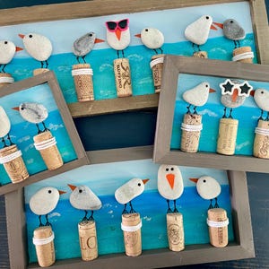 May include: Handmade framed artwork featuring whimsical birds crafted from pebbles, perched atop wine corks. Each bird has a unique expression, some with sunglasses. The background depicts a blue ocean scene, creating a coastal theme.