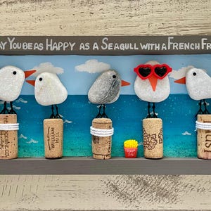 May include: A rectangular wall art piece in a gray frame featuring five pebble seagulls perched on wine corks against a painted blue ocean background. The text reads, May You Be As Happy As A Seagull With A French Fry! One seagull wears red heart-shaped glasses.