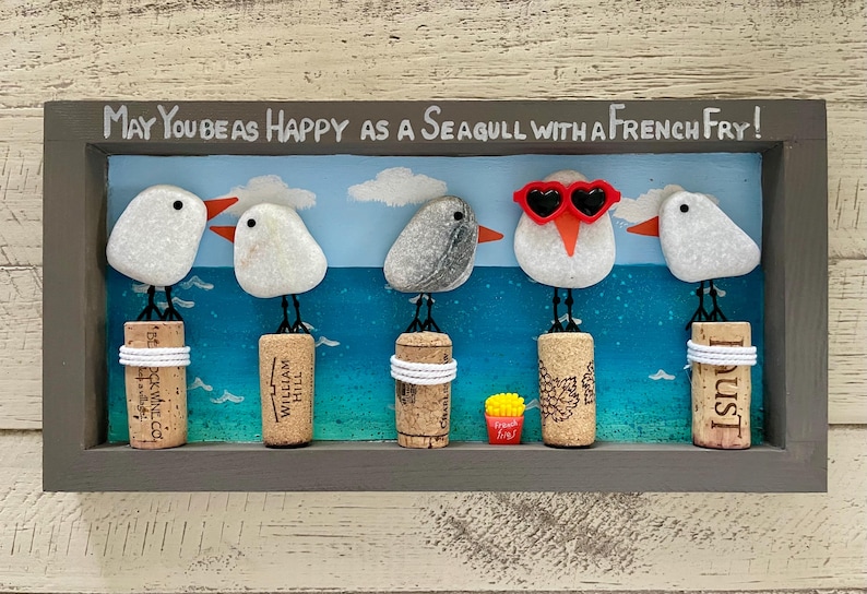 May include: A rectangular wall art piece in a gray frame featuring five pebble seagulls perched on wine corks against a painted blue ocean background. The text reads, May You Be As Happy As A Seagull With A French Fry! One seagull wears red heart-shaped glasses.