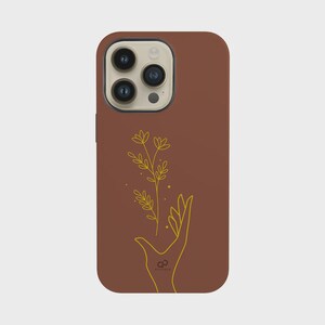 May include: A brown phone case featuring a gold outline of a hand holding a plant with leaves and flowers. The phone case has a smooth finish and a camera cutout at the top.