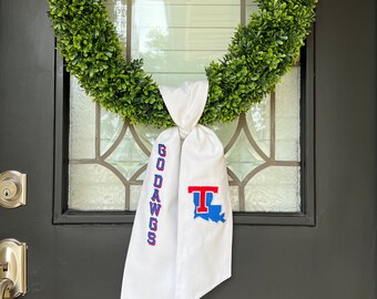 May include: A white door wreath with a green artificial boxwood wreath and a white fabric bow with the text "Go Dawgs" and a red and blue Louisiana State University logo.