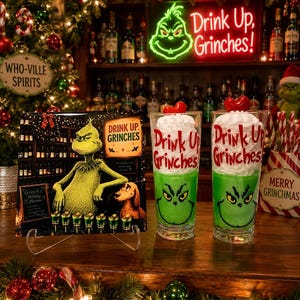 Könnte beinhalten: Zwei klare Trinkgläser gefüllt mit grüner Flüssigkeit, weißer Schlagsahne und einer roten herzförmigen Garnitur. Die Gläser zeigen den Text Drink Up Grinches und eine grüne Gesichtsgrafik. Ein passendes quadratisches Schild zeigt das gleiche Design.