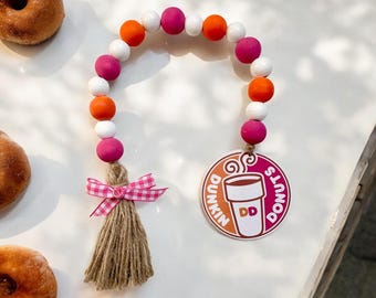 Può includere: Una ghirlanda di perline rosa, arancio e bianche con un fiocco di pizzo rosa e bianco e un pon pon marrone. La ghirlanda ha un'etichetta rotonda di legno con il logo Dunkin' Donuts e il testo "Dunkin' Donuts".