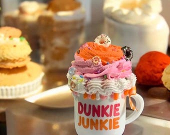 Może przedstawiać: Biały kubek z napisem "DUNKI JUNKIE" w różowych i pomarańczowych literach. Kubek zwieńczony jest kolorowym, mrożonym deserem z mini pączkiem na wierzchu. Inne desery są w tle.