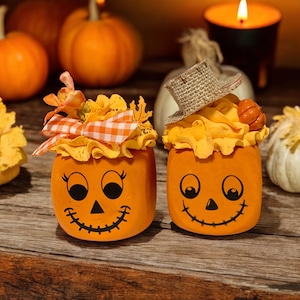 May include: Two orange pumpkin-shaped containers with painted faces. One container has a brown and white straw hat on top. Both containers have a black painted smile and eyes.