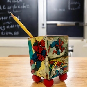 Peut inclure: Un porte-crayons en forme de boîte à roulettes, décoré avec des images de Spider-Man tirées d'une bande dessinée. Le porte-crayons est rouge et bleu et repose sur une surface en bois.
