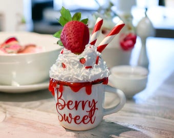 May include: A white ceramic mug with a red rim and the words "Berry Sweet" in red script. The mug is topped with whipped cream, a strawberry, and candy cane sticks. Red sprinkles and strawberry accents complete the design.