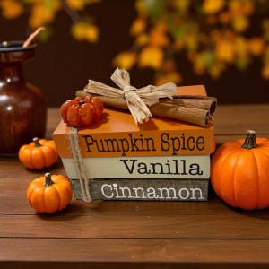 May include: A stack of decorative books with the words "Pumpkin Spice", "Vanilla", and "Cinnamon" printed on them. The books are orange, cream, and gray. Cinnamon sticks and a small pumpkin are on top. Several small orange pumpkins are scattered around.