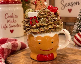 May include: A ceramic mug shaped like a gingerbread person with a red bow tie, topped with faux whipped cream, a gingerbread cookie figure, a red heart, and a small stocking ornament. The mug features a smiling face and a white handle with colorful sprinkles.