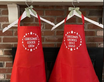 Könnte beinhalten: Zwei rote Schürzen mit weißem Text und weihnachtlichen Grafiken. Eine Schürze sagt "Christmas Cookie Crew" und die andere sagt "Christmas Calories Don't Count".