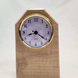 May include: A wooden clock with a light wood finish and a white face with black numbers and hands. The clock is labeled "QUARTZ" and has a gold trim.