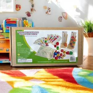 May include: A box for a wooden toy set called "Jumbo Sequence Stacker" by Constructive Playthings. The box shows the contents of the set, including colorful wooden rings, a wooden stacking tower, and cards with images of the rings. The box is brown with white text and a green drawstring bag.