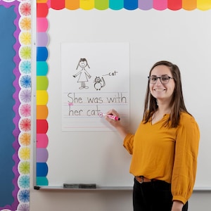 May include: A white paper with a drawing of a girl and a cat, with the text "She was with her cat." A person is holding a pink marker and writing on the paper. The background is a white board with colorful decorations.