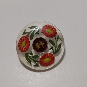 May include: A clear glass button with a floral design. The button has three red flowers with yellow centers and green leaves.