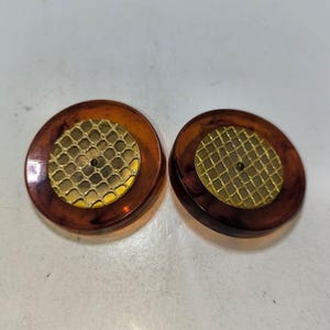 Puede incluir: Un par de pendientes redondos, marrones y dorados con un patrón geométrico. Los pendientes están hechos de plástico y tienen una inserción de metal dorado.