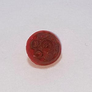 May include: A small, round, red button with a detailed, embossed floral design. The button is a solid, vibrant red color and appears to be made of a smooth, possibly glass-like material. The floral pattern is visible in the center.