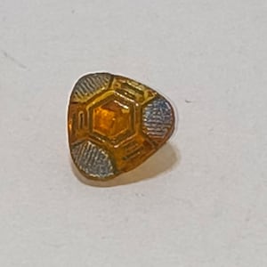 May include: A small, triangular pin with a geometric design. The pin features a central orange hexagon surrounded by gold and silver-colored segments. The silver segments have a textured, ridged pattern.