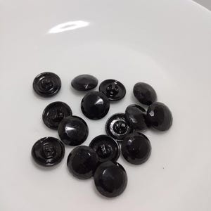 Może przedstawiać: Kolekcja fasetowanych, czarnych guzików z błyszczącym wykończeniem. Guziki są okrągłe, lekko wypukłe i mają trzpień z tyłu do przyszywania. Rozrzucone na białej powierzchni.