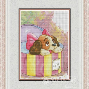 Könnte beinhalten: Ein Kreuzstichmuster eines braunen und weißen Welpen mit einer rosa Schleife, der aus einer rosa und gelb gestreiften Geschenkbox mit dem Wort "Lady" auf einem Etikett schaut.