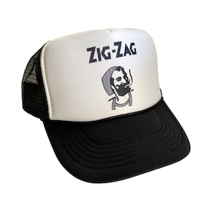 May include: White trucker hat with a black mesh back. The front of the hat has a black and white illustration of a man smoking a cigarette with the text "ZIG-ZAG" above him.