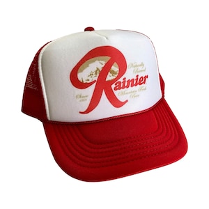 May include: A red and white trucker hat with a red "R" logo and the words "Naturally Brewed Rainier Mountain Fresh Beer" in gold lettering. The hat has a mesh back.