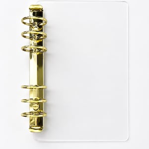 May include: A gold-colored binder mechanism with four rings is attached to a clear, rectangular sheet of plastic. The binder is made of shiny metal and is mounted on the left side of the plastic sheet. The background is white.