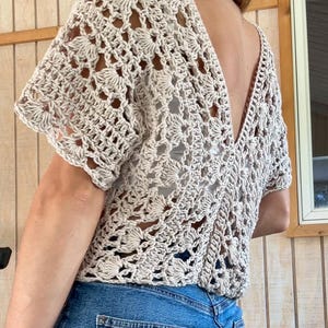 May include: A beige crochet top with a deep V-neck and short sleeves. The openwork design features floral patterns. The top is paired with blue denim jeans.