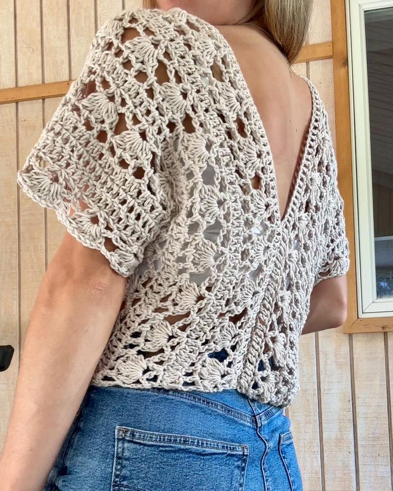 May include: A beige crochet top with a deep V-neck and short sleeves. The openwork design features floral patterns. The top is paired with blue denim jeans.