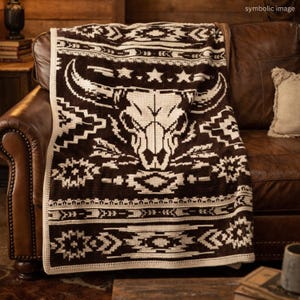 May include: A brown and cream-colored throw blanket with a Southwestern design featuring a bull skull and star pattern. The blanket is draped over a brown leather sofa, showcasing its intricate crochet work.