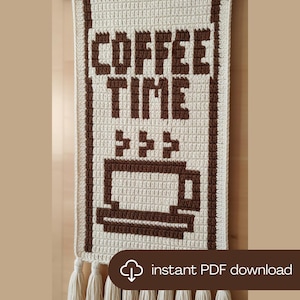 Op de afbeelding: Gehaakte wandhanger in crème met de tekst "COFFEE TIME" in bruin. Een bruine koffiekopjesafbeelding staat eronder. De wandhanger heeft een rand en kwasten aan de onderkant. De tekst "instant PDF download" is ook zichtbaar.