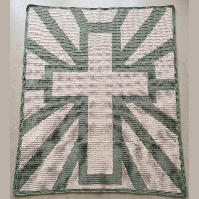 May include: A square, handmade crochet blanket featuring a large cross design. The cross and radiating lines are sage green, set against a cream-colored background. The blanket has a sage green border.
