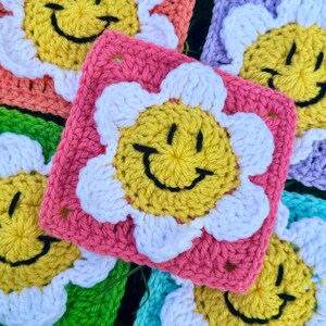 May include: Close-up of several colourful crochet squares. Each square features a yellow smiley face with a black outline, surrounded by white petals. The squares are in various colours, including pink, orange, green, and purple. The squares are handmade.