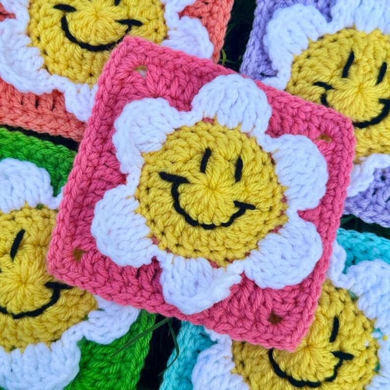 May include: Close-up of several colourful crochet squares. Each square features a yellow smiley face with a black outline, surrounded by white petals. The squares are in various colours, including pink, orange, green, and purple. The squares are handmade.